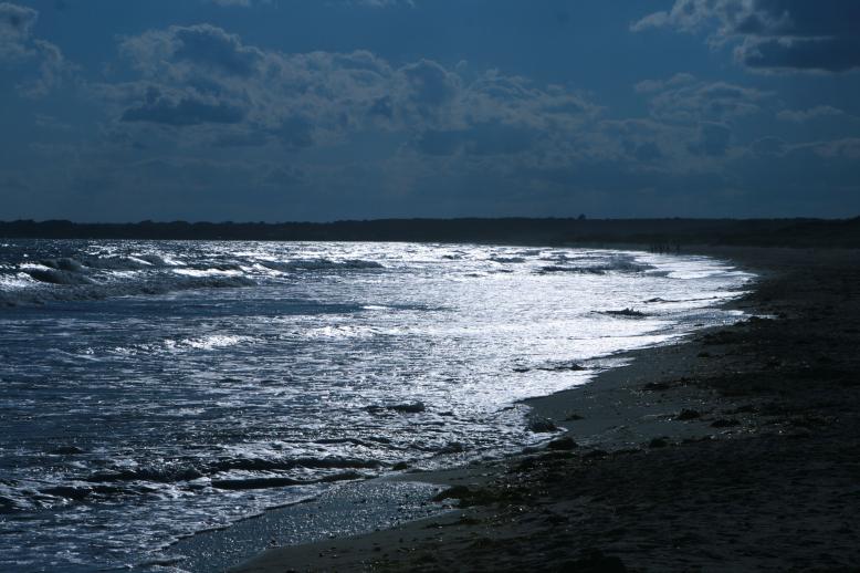 The ocean waves meet the beach which sweeps in a long arc.  The late day sun provides a reflection on the breaking waves that produces and intense image of bright light against a sweeping sea. Horseneck Beach, 6:30 PM, July 4, 2009.