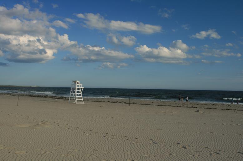 Soft puffs of clouds above the sea.  There are people standing near the shore, obscured, you can't tell who they are.  There are only a few.  The sun is behind a cloud but the far away ocean is in full sun.  An empty lifeguard chair is prominent on the left side of this photo.
