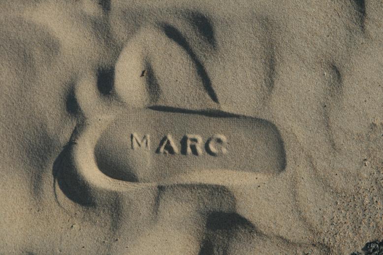 A childs sneaker print in the sand.  The word 'MARC' is embossed into the sole of the shoe and promonent in the center of the shoe print.